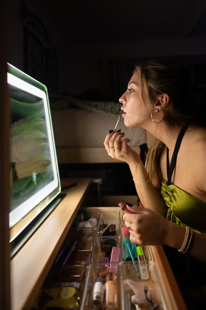 A girl gets ready by putting a makeup product on her lips in a mirror.