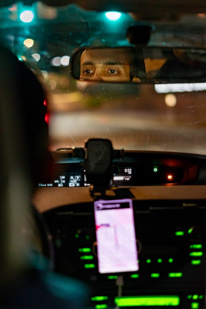 A man drives at night, his eyes can be seen through the rearview mirror of his car.